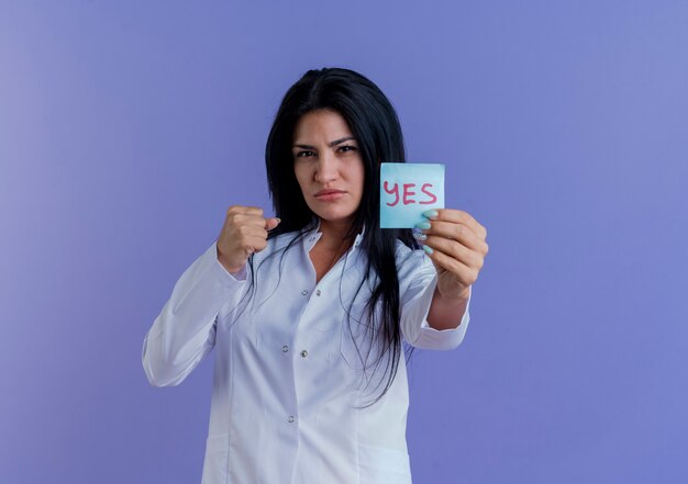 Confident young female doctor wearing medical robe showing yes note looking clenching fist isolated