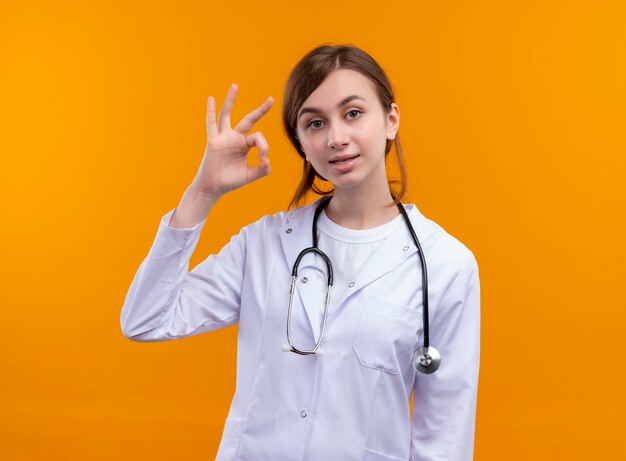 Confident young female doctor wearing medical robe and stethoscope doing ok sign on isolated orange space