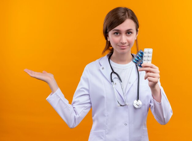 의료 가운과 청진기를 착용하고 의료 약품을 들고 고립 된 오렌지 공간에 빈 손을 보여주는 확신 젊은 여성 의사