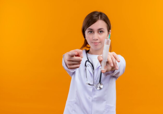 Confident young female doctor wearing medical robe and stethoscope holding syringe and pointing  on isolated orange space with copy space
