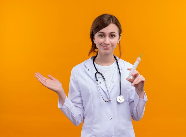 Confident young female doctor wearing medical robe and stethoscope holding syringe and showing empty hand on isolated orange space