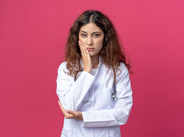 Confident young female doctor wearing medical robe and stethoscope keeping hand on elbow and on face isolated on pink wall with copy space