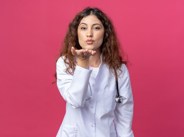 Confident young female doctor wearing medical robe and stethoscope sending blow kiss isolated on pink wall with copy space