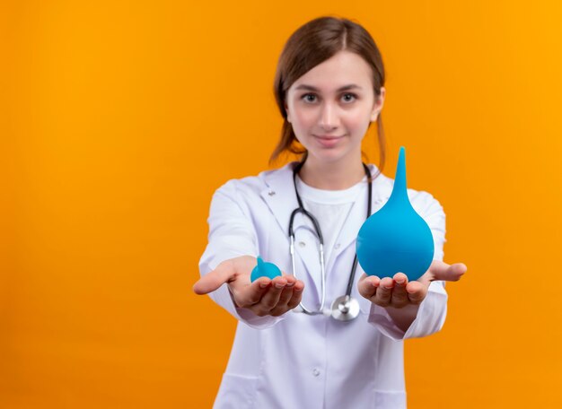 Confident young female doctor wearing medical robe and stethoscope stretching enemas on isolated orange space with copy space