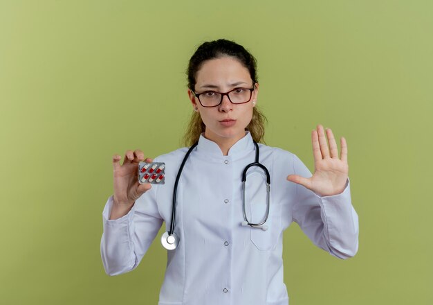 Confident young female doctor wearing medical robe and stethoscope with glasses holding pills showing stop gesture isolated on olive green wall