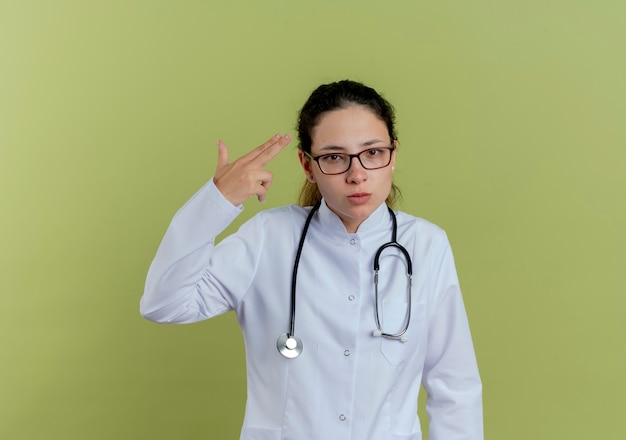 Confident young female doctor wearing medical robe and stethoscope with glasses showing pistol gesture isolated on olive green wall
