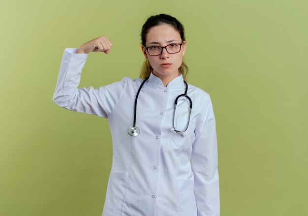 Confident young female doctor wearing medical robe and stethoscope with glasses showing strong gesture isolated on olive green wall