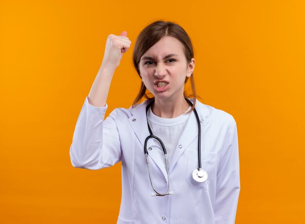 Уверенная молодая женщина-врач в медицинском халате и стетоскопе с поднятым кулаком на изолированном оранжевом пространстве с копией пространства