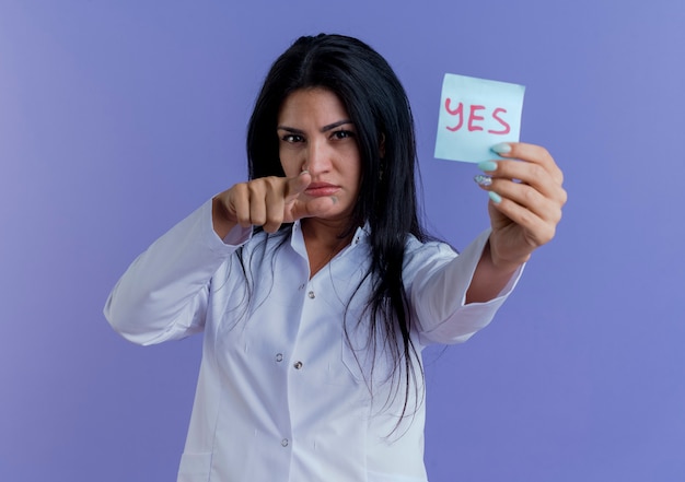 Confident young female doctor wearing medical robe stretching out yes note, looking and pointing 