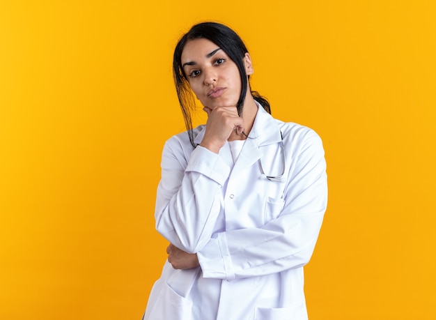 Confident young female doctor wearing medical robe with stethoscope grabbed chin isolated on yellow wall