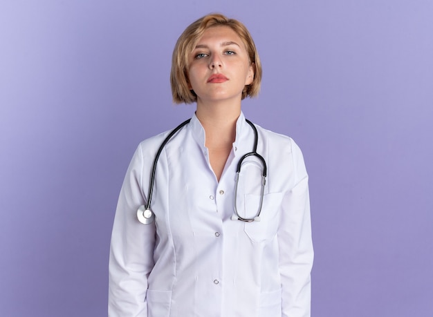 Confident young female doctor wearing medical robe with stethoscope isolated on blue wall