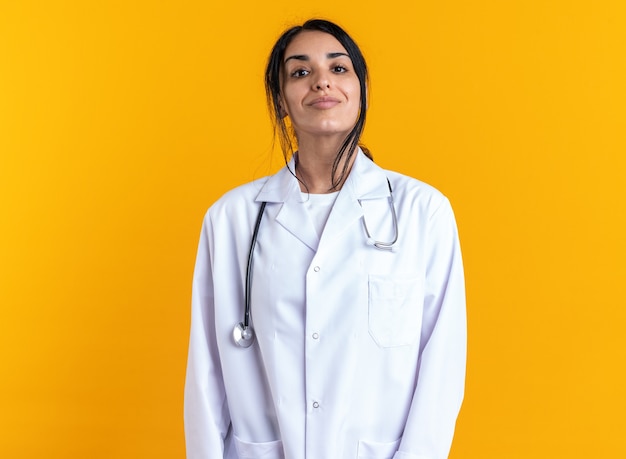 Confident young female doctor wearing medical robe with stethoscope isolated on yellow wall