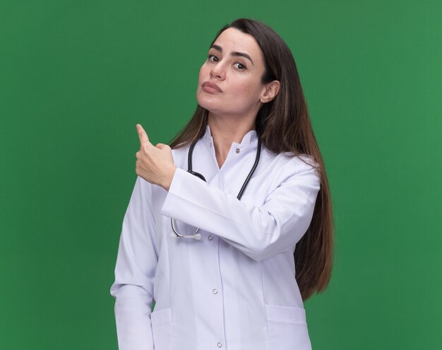 Confident young female doctor wearing medical robe with stethoscope points back on green 