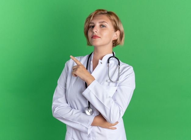 Confident young female doctor wearing medical robe with stethoscope points at side isolated on green wall with copy space