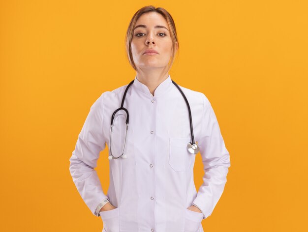 Confident young female doctor wearing medical robe with stethoscope putting hands on pocket isolated on yellow wall