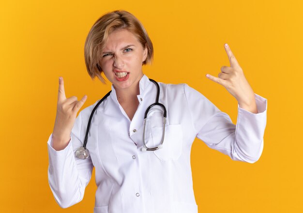 Confident young female doctor wearing medical robe with stethoscope showing goat gesture isolated on orange wall