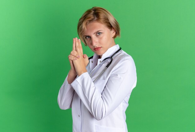 Confident young female doctor wearing medical robe with stethoscope showing pistol gesture isolated on green wall