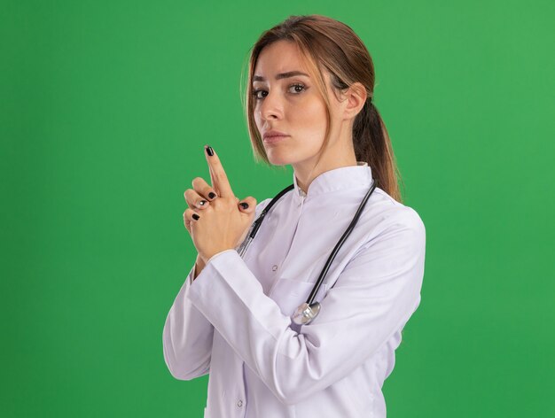緑の壁に分離されたピストルジェスチャーを示す聴診器で医療ローブを身に着けている自信を持って若い女性医師