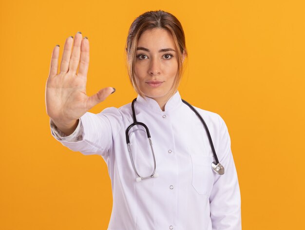 Confident young female doctor wearing medical robe with stethoscope showing stop gesture isolated on yellow wall