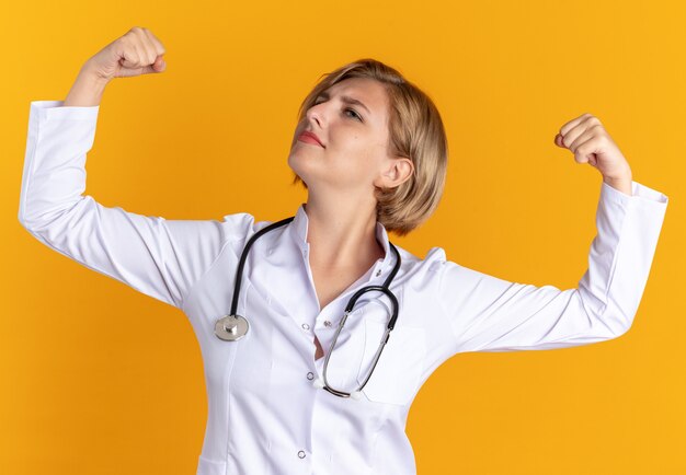 Уверенная молодая женщина-врач в медицинском халате со стетоскопом показывает сильный жест, изолированный на оранжевой стене