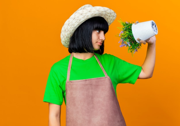 無料写真 ガーデニング帽子をかぶった制服を着た自信を持って若い女性の庭師は、植木鉢の花を保持し、見ています