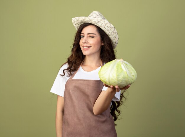 Confident young female gardener in uniform wearing gardening hat blinks eye and holds cabbage isolated on olive green wall