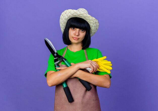 Уверенная молодая женщина-садовник в униформе в садовой шляпе скрещивает руки с ножницами и перчатками