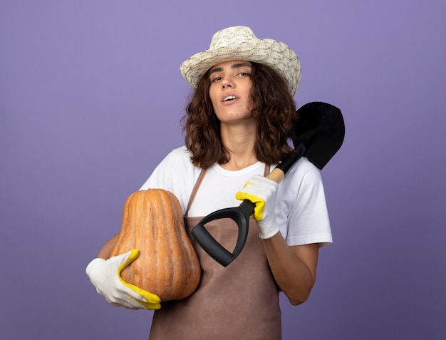 Confident young female gardener in uniform wearing gardening hat and gloves holding pumpkin putting spade on shoulder