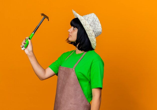 Free photo confident young female gardener in uniform wearing gardening hat holding hoe rake