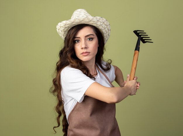 Confident young female gardener in uniform wearing gardening hat holding rake isolated on olive green wall