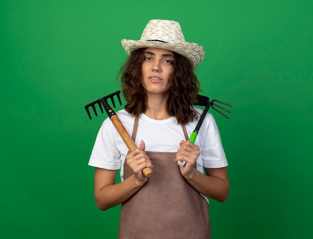 Confident young female gardener in uniform wearing gardening hat holding rake with hoe rake on shoulders