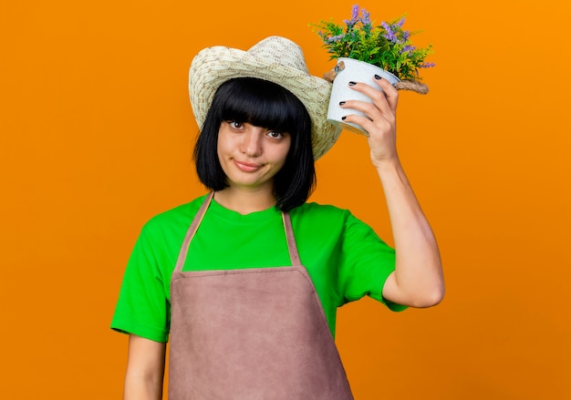 Confident young female gardener in uniform wearing gardening hat holds flowerpot
