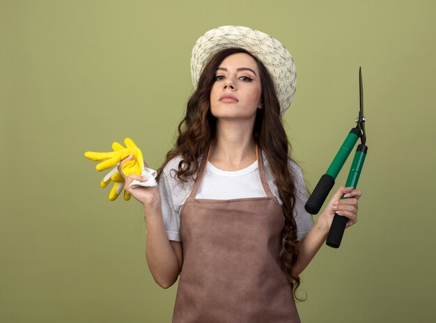 Confident young female gardener in uniform wearing gardening hat holds garden clippers and gloves isolated on olive green wall