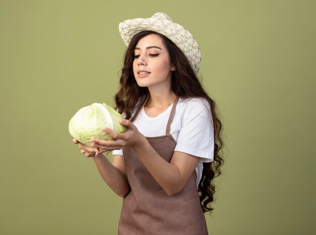 Confident young female gardener in uniform wearing gardening hat holds and looks at cabbage isolated on olive green wall