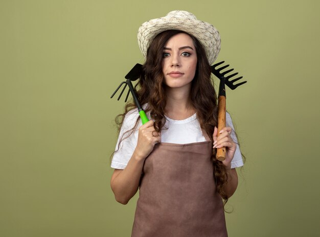 Уверенная молодая женщина-садовник в униформе в садовой шляпе держит грабли и мотыгу на плечах, изолированные на оливково-зеленой стене