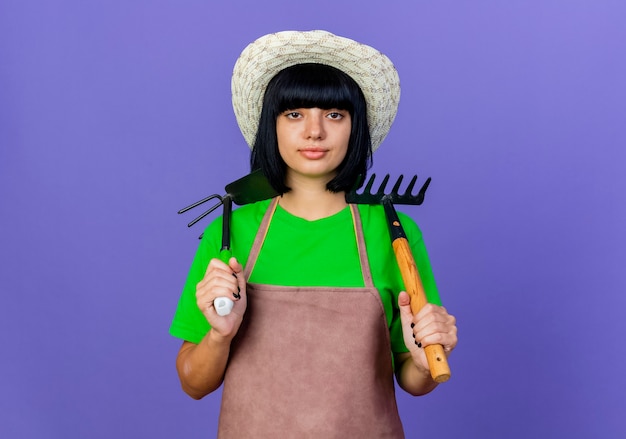 Free photo confident young female gardener in uniform wearing gardening hat holds rake and hoe rake