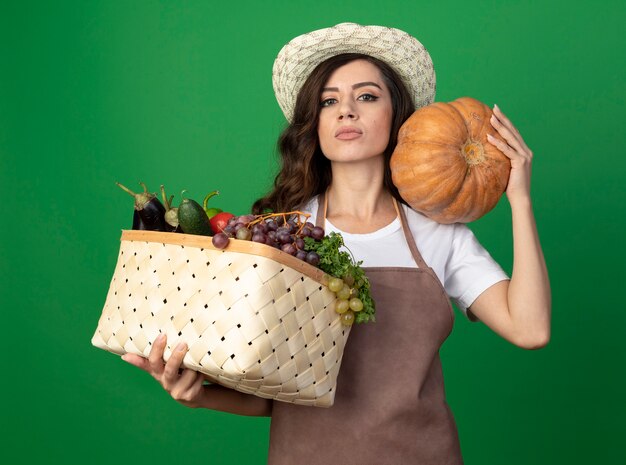 Уверенная молодая женщина-садовник в униформе в садовой шляпе держит корзину с овощами и тыкву на плече, изолированную на зеленой стене с копией пространства