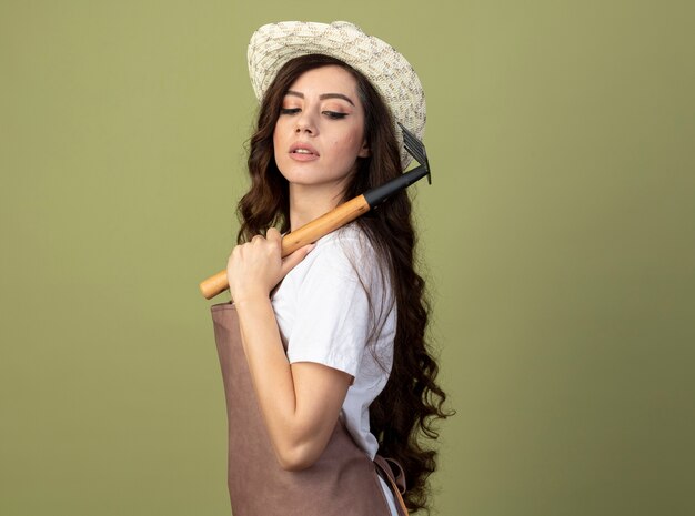 Fiducioso giovane giardiniere femminile in uniforme che indossa il cappello da giardinaggio si trova lateralmente tenendo il rastrello sulla spalla isolato sulla parete verde oliva