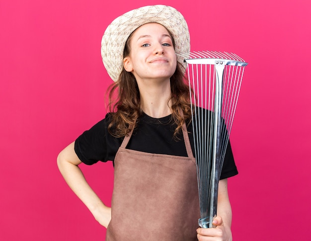 Confident young female gardener wearing gardening hat holding leaf rake putting hand on hip