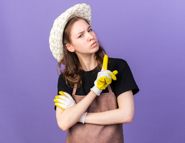 青い壁に隔離されたものを示す手袋とガーデニング帽子を身に着けている自信を持って若い女性の庭師