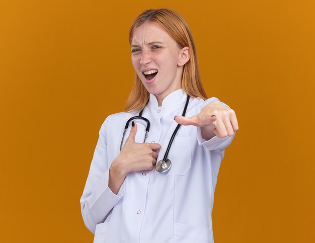 Уверенная молодая женщина-врач-имбирь в медицинском халате и стетоскопе смотрит и указывает пальцем вверх, изолирована на оранжевой стене с копией пространства