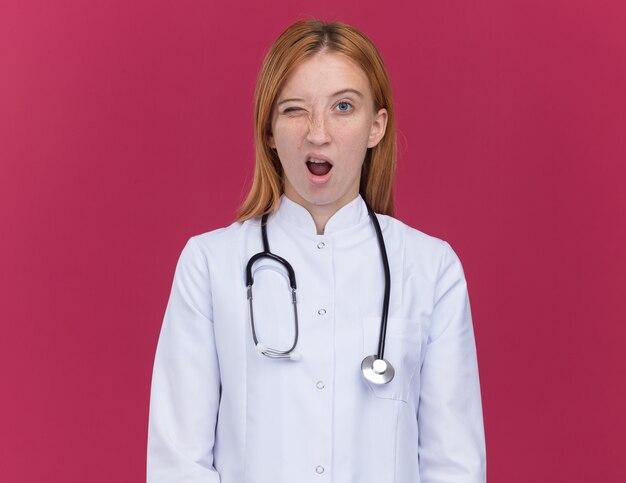 Уверенная молодая женщина-рыжий врач в медицинском халате и подмигивает стетоскопом, изолированным на малиновой стене