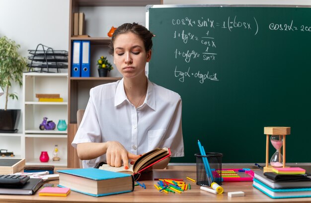 уверенная в себе молодая учительница математики сидит за столом со школьными принадлежностями, указывая пальцем на открытую книгу и глядя на нее в классе