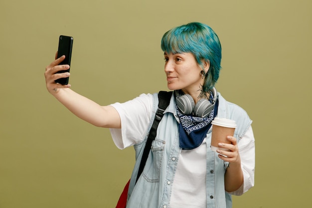 Confident young female student wearing headphones and bandana on neck and backpack holding paper coffee cup taking selfie with mobile phone isolated on olive green background