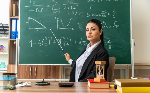 confident young female teacher sits at table with school supplies points at blackboard with pointer stick in classroom