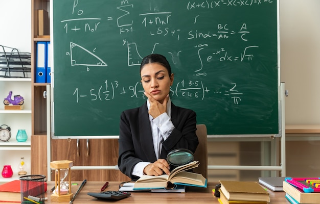 Confident young female teacher sits at table with school tools reading book with magnifier grabbed chin in classroom