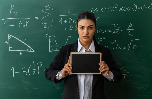 confident young female teacher standing in front blackboard holding mini blackboard in classroom