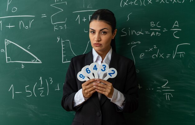 confident young female teacher standing in front blackboard holding number fans in classroom