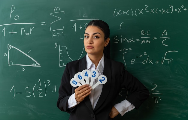 confident young female teacher standing in front blackboard holding number fans putting hand on board in classroom