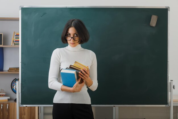 confident young female teacher wearing glasses holding books standing in front blackboard in classroom